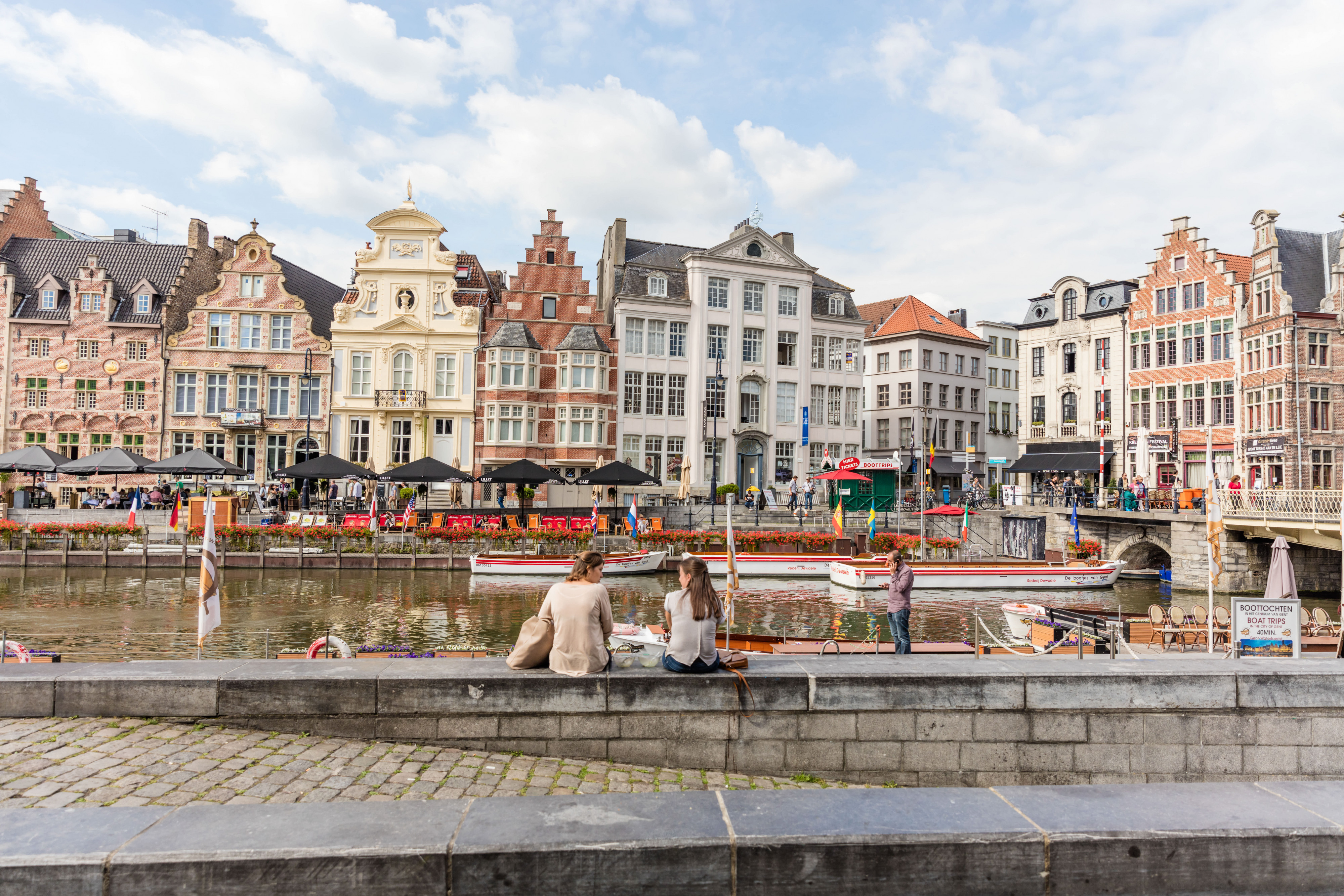 Canalfront cafes and historic gabled buildings People sit by a canal with tour boats and outdoor cafes, backed by historic gabled buildings under a blue sky.