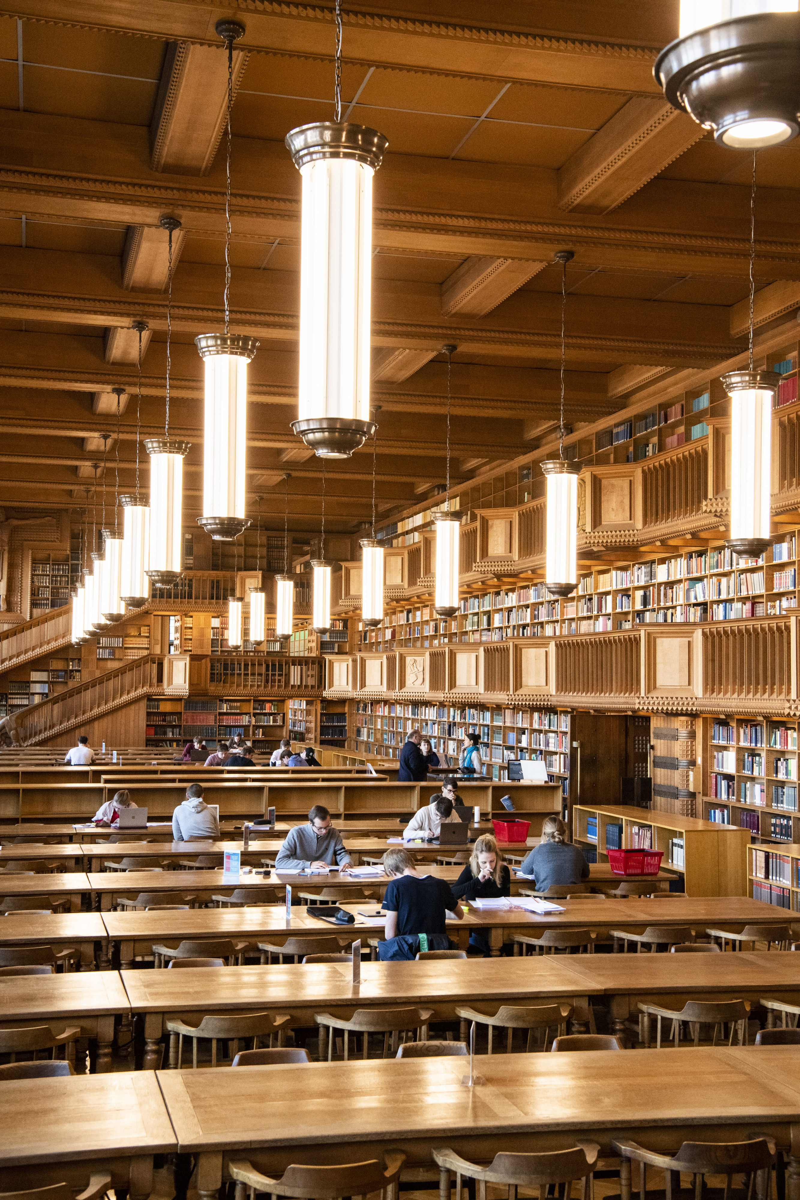 Library reading room with hanging lights Wood-paneled library reading room with tall hanging lights, long tables, bookshelves, and students studying