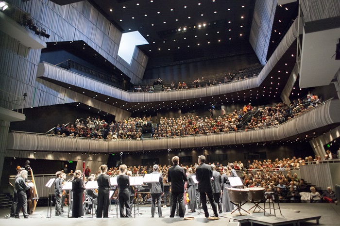 Orchestra Performing in a Packed Concert Hall Orchestra and conductor on stage facing a full, multi-level concert hall audience.