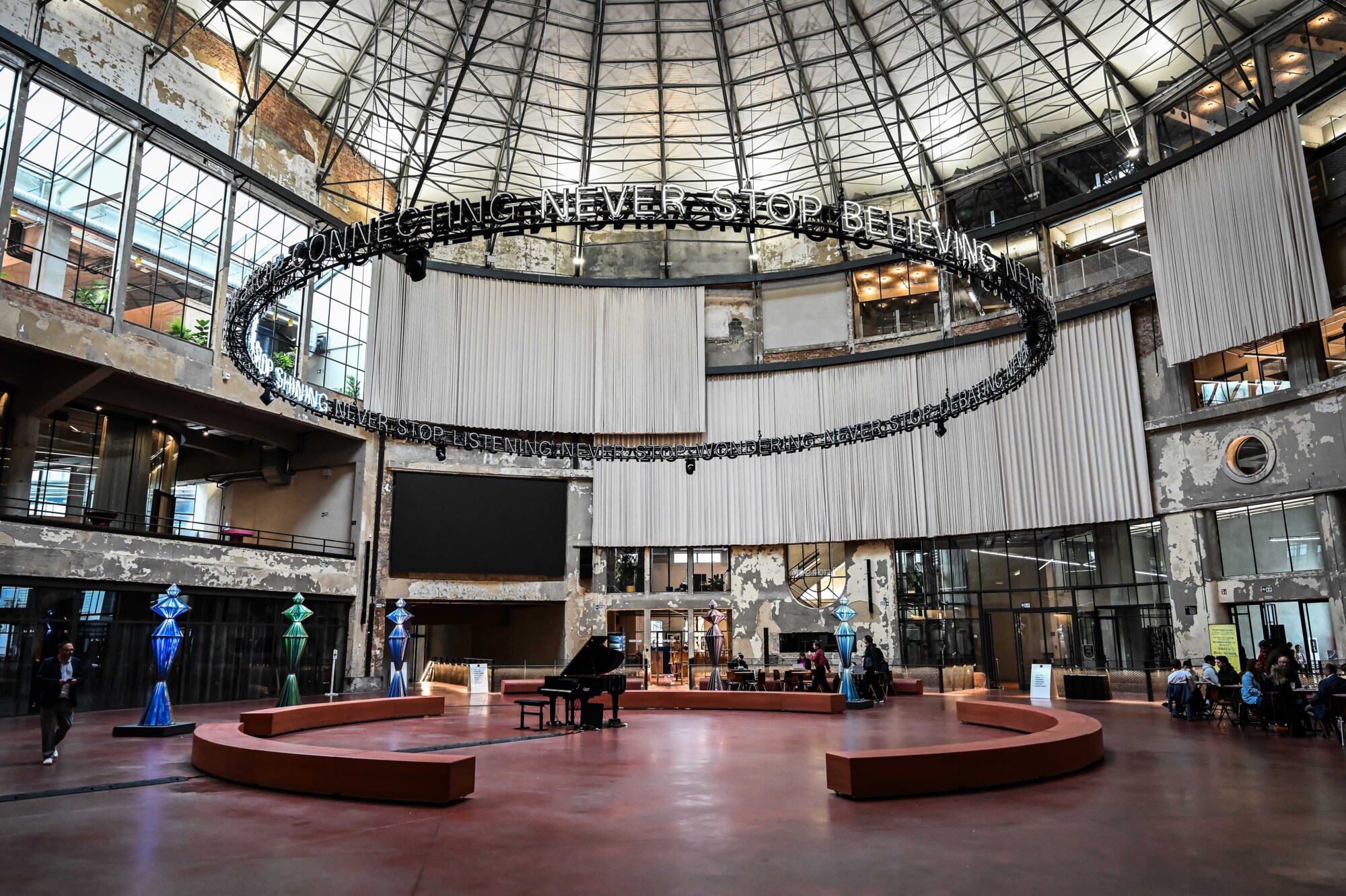 Industrial atrium with hanging text sculpture Industrial atrium with glass roof, circular hanging text sculpture, benches and piano, and people seated at tables.