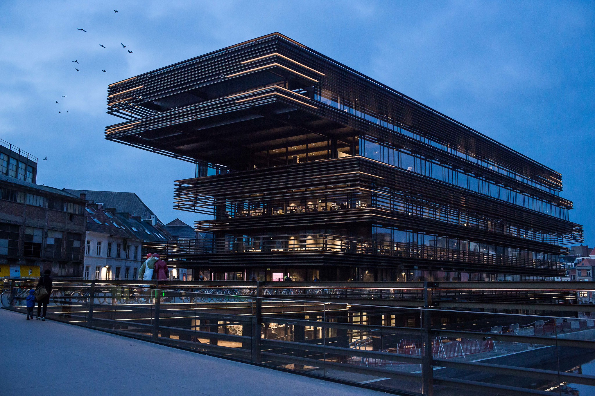 Modern waterfront building at dusk Modern glass-and-metal building lit at dusk beside a canal, with pedestrians on a bridge and birds in the sky.