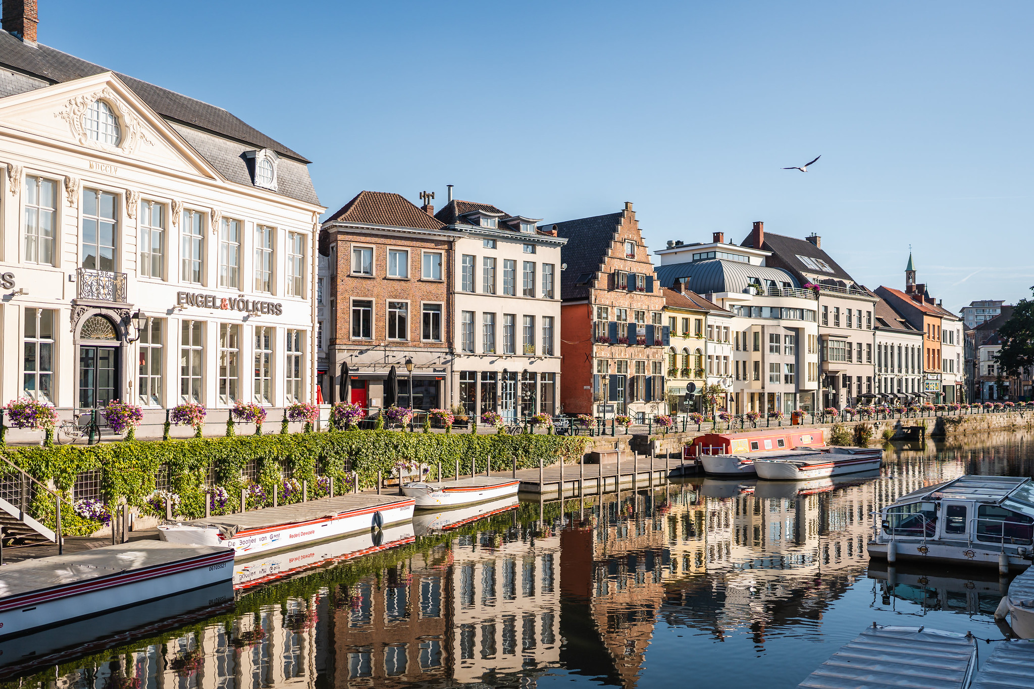 Historic canal waterfront with boats Canal with moored boats and historic waterfront buildings reflected in calm water under a clear blue sky.
