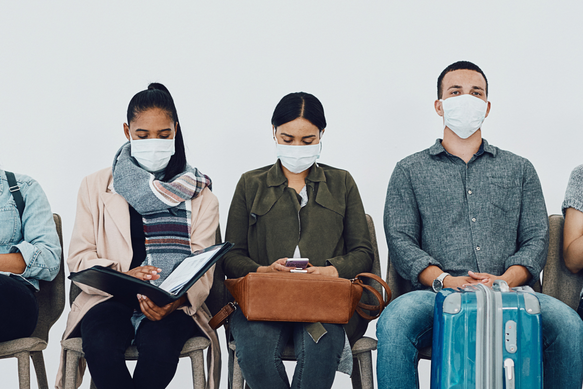 Five travelers wearing face masks sit in a row with luggage, looking at phones and documents.