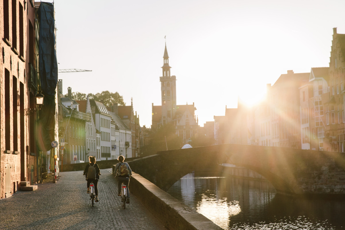 Cyclists by canal at sunset Two cyclists ride along a canal path at sunset, with a stone bridge and a tall clock tower in the distance.