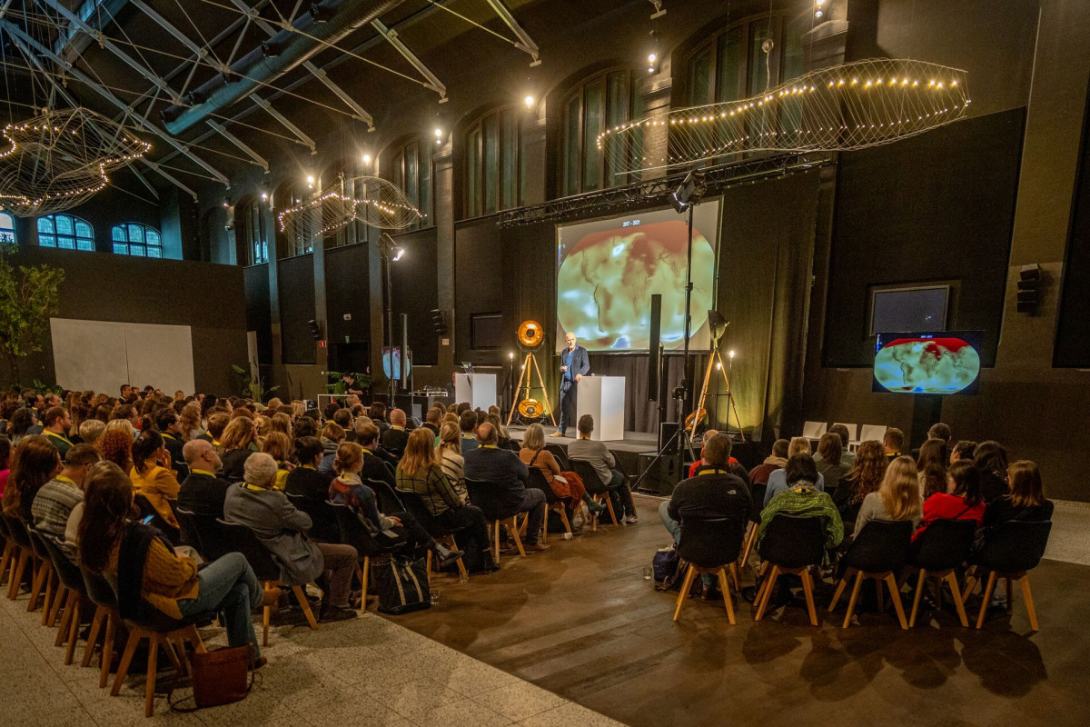 Conference talk with projected world map Audience seated in a large hall watching a speaker on stage beside a projected world map.