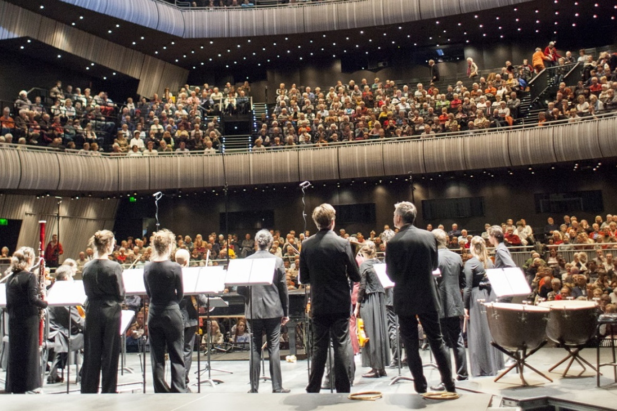 Orchestra performing to a full concert hall Orchestra on stage facing a packed concert hall audience, viewed from behind the musicians.