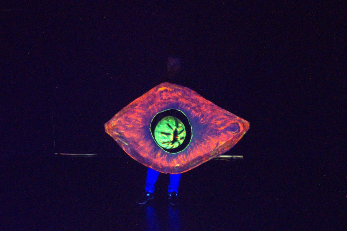 Glowing Eye Costume on Dark Stage Person in dark wearing a glowing eye-shaped costume with a green iris and red-orange outline on a black stage