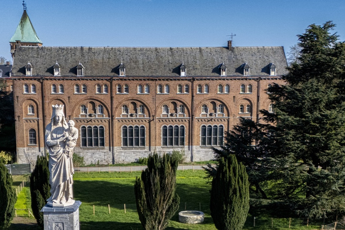 Statue of the Virgin Mary holding a child in front of a large brick monastery-like building, surrounded by trees.